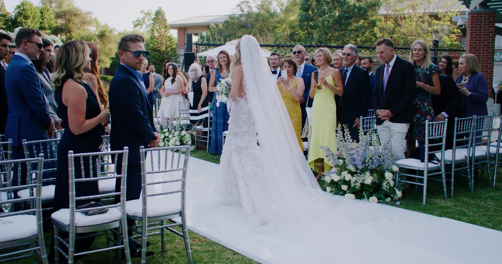 Alla walking the aisle with a long veil as guests stand, in the gardens at Four Seasons Westlake Village