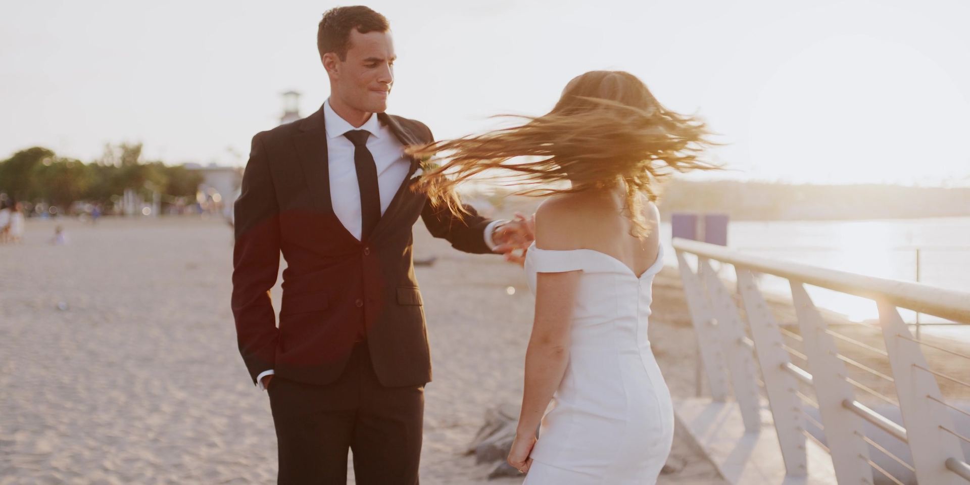Alex and Anna on the sand, her hair blowing across her face, water and low sun behind.