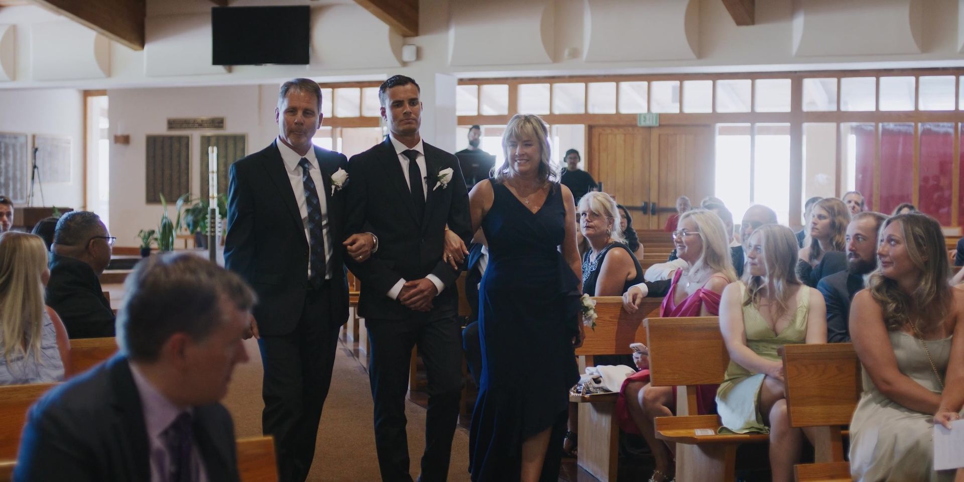 The groom walking down the church aisle arm in arm with a man and a woman, guests seated in the pews on both sides.