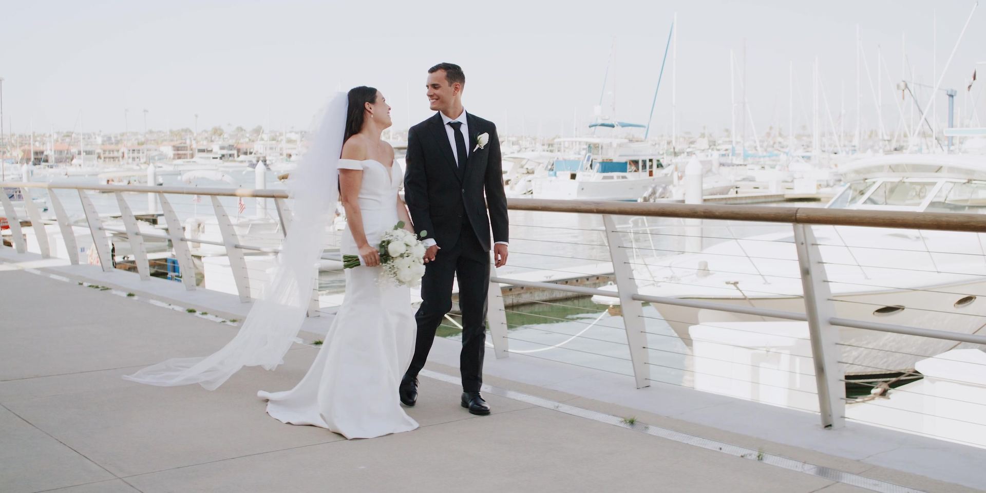 Alex and Anna at the rail of the marina walkway, boats moored along the slips behind them.