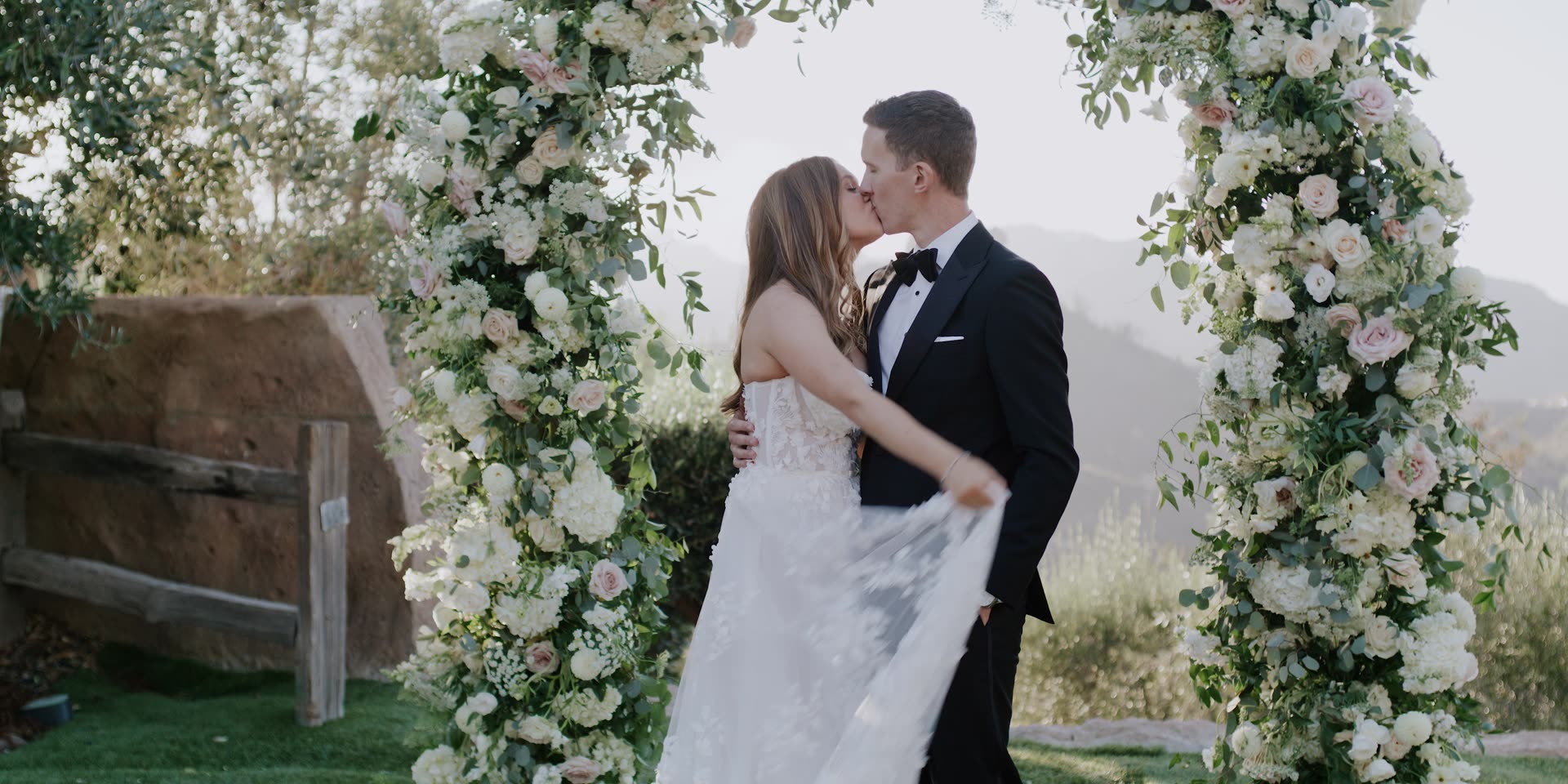 Andy and Emily kissing beneath the floral arch at Cielo Farms with the Malibu hills behind