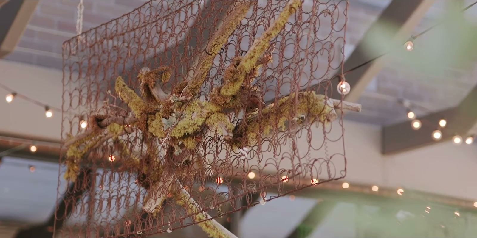 A rusted bedspring frame woven with moss-covered branches, hung under string lights.