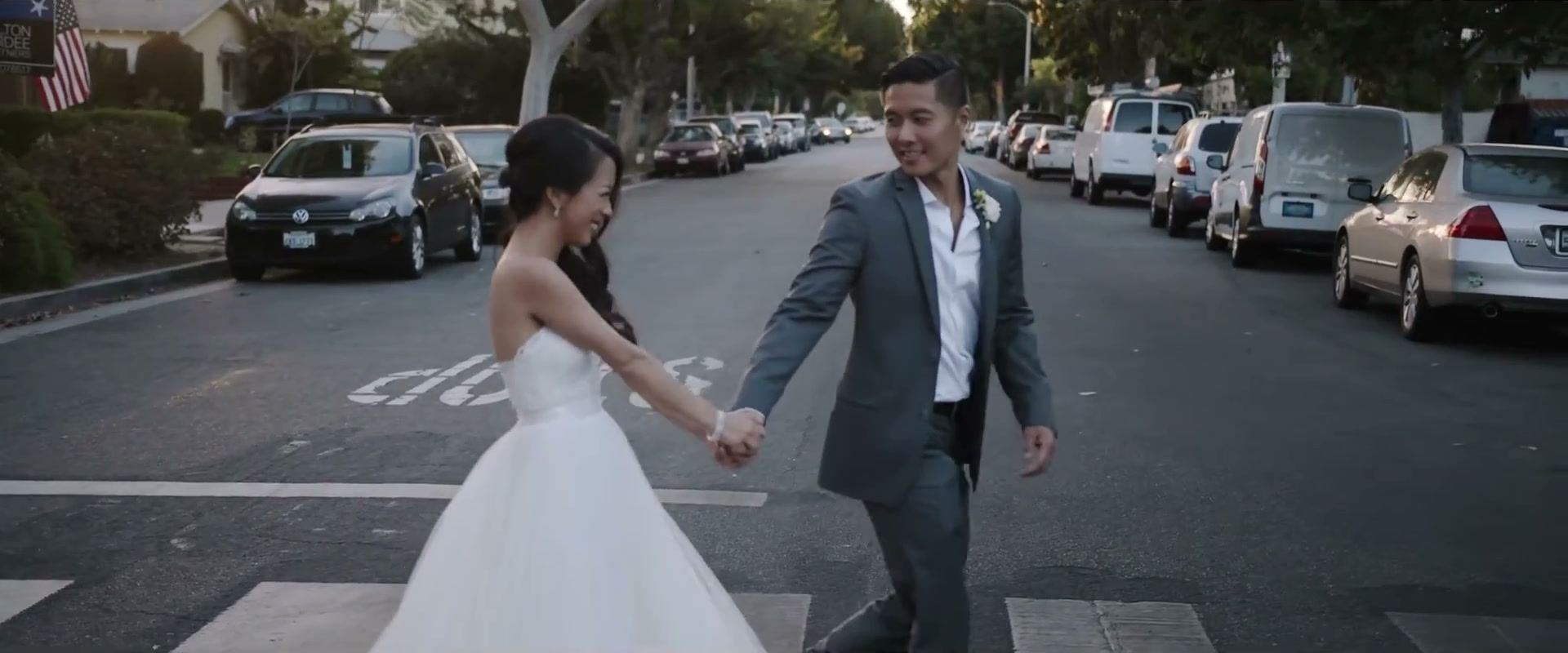 Andrea and Gene crossing a quiet street hand in hand, parked cars along both sides.