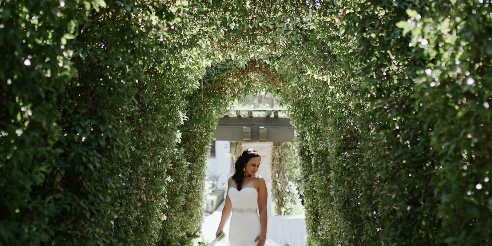 A bride stepping out of a long arched tunnel of green toward the camera.