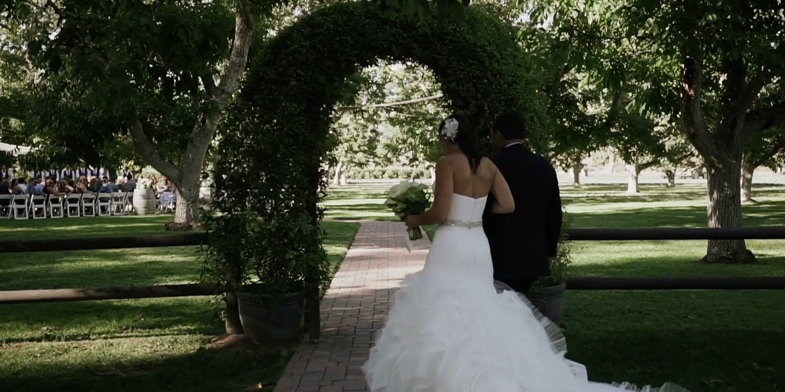 A bride walking along a brick path toward the green arch, rows of white chairs beyond it in the grove.