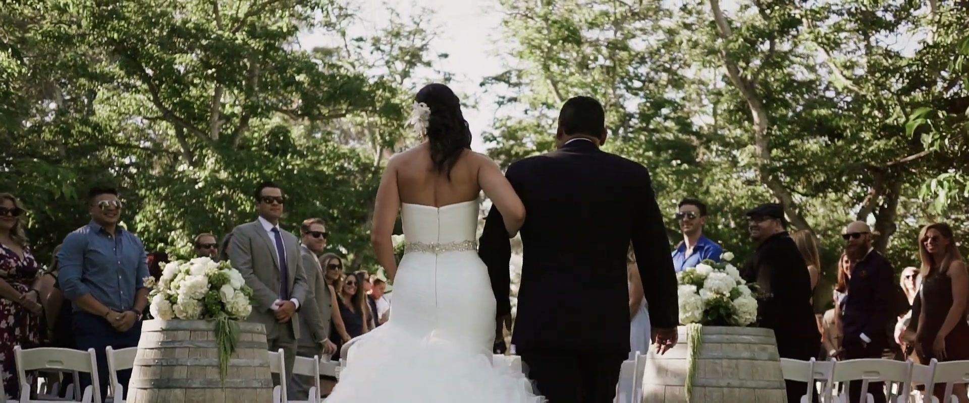 Lindsay walking down the aisle on the arm of a man in a dark suit, between standing guests and barrels topped with white hydrangea.