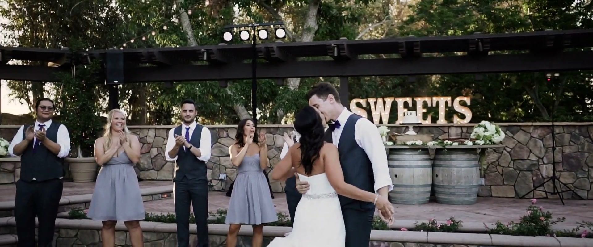 Alex and Lindsay kissing mid-dance on the stone-edged floor, bridesmaids and groomsmen clapping, a SWEETS marquee sign behind.