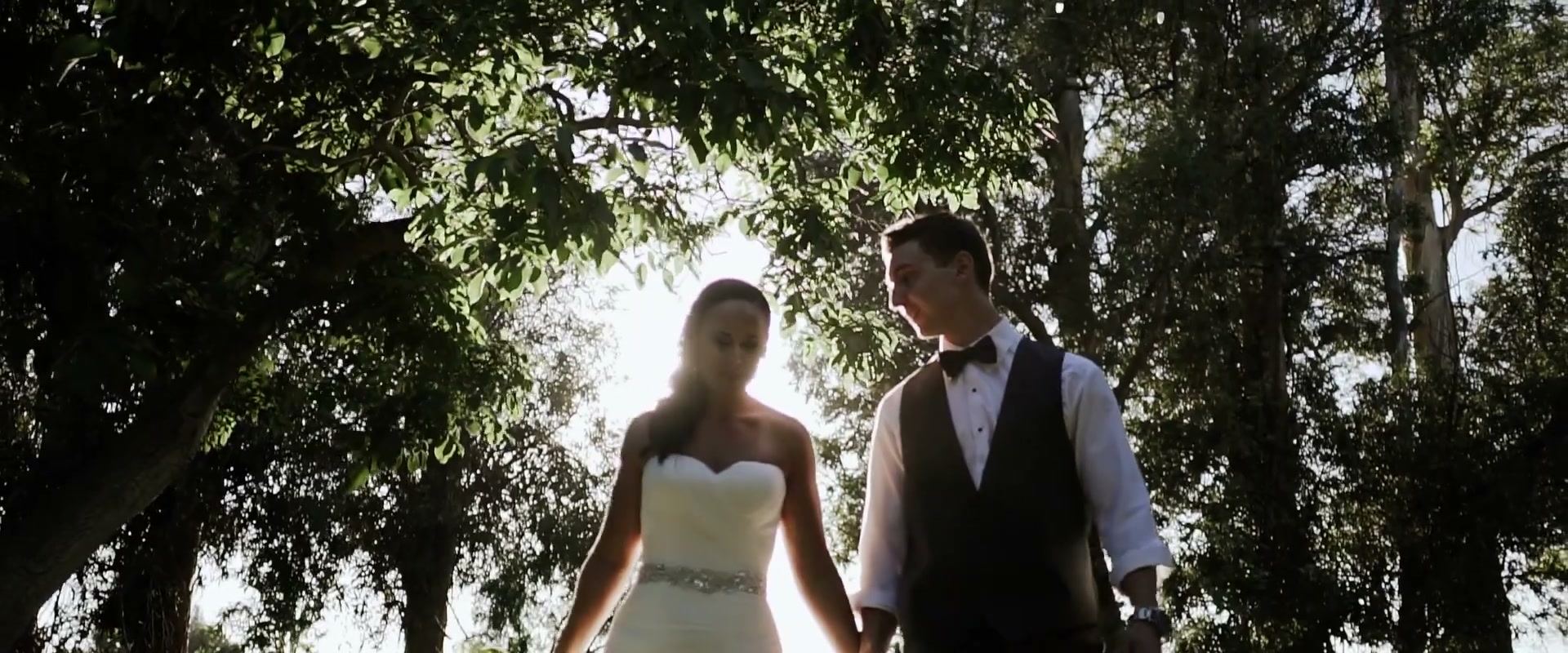 Alex and Lindsay walking toward the camera under dark walnut branches, bright light behind them.