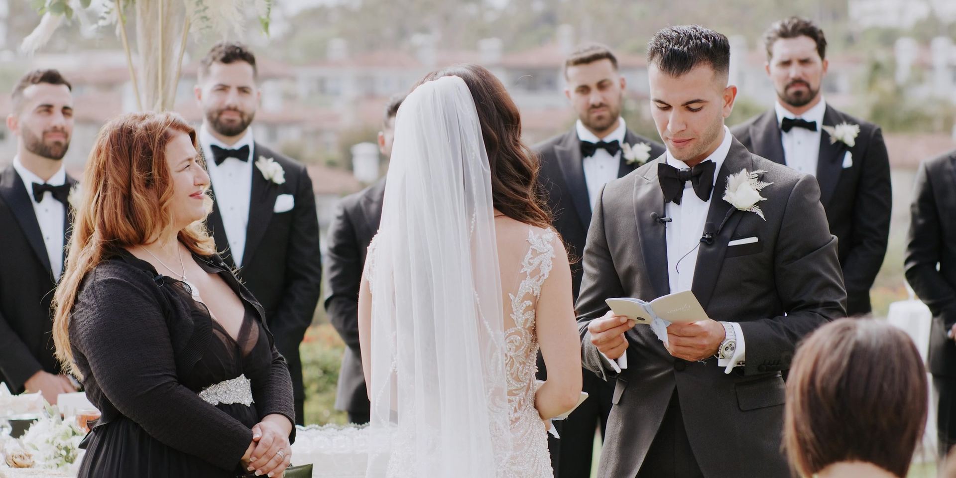 Aurash reading his vows from a folded card, Madison facing him under her veil, the wedding party in black tie behind them.