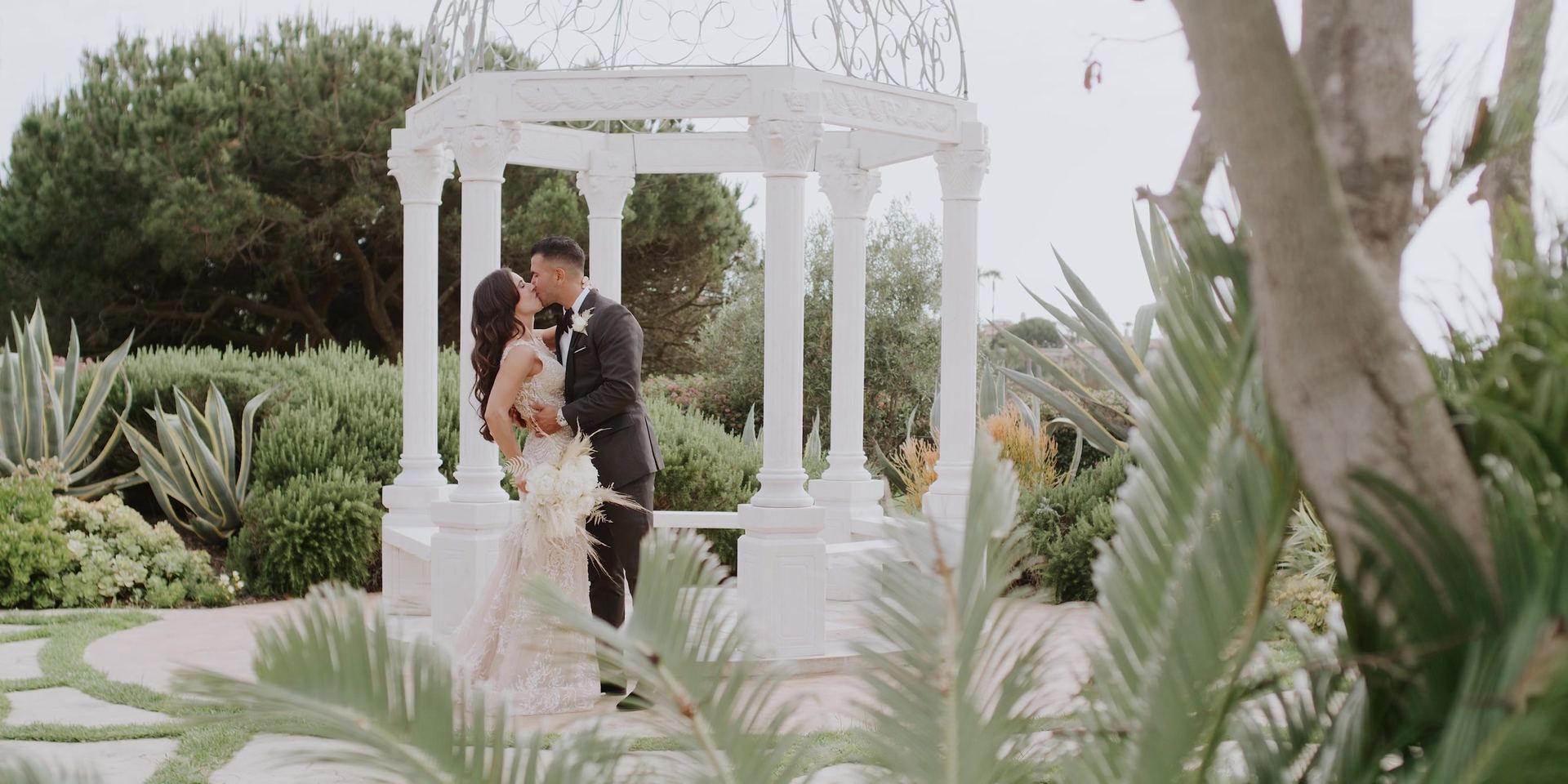 Aurash and Madison kissing in front of the white rotunda, framed by palm fronds and agave.