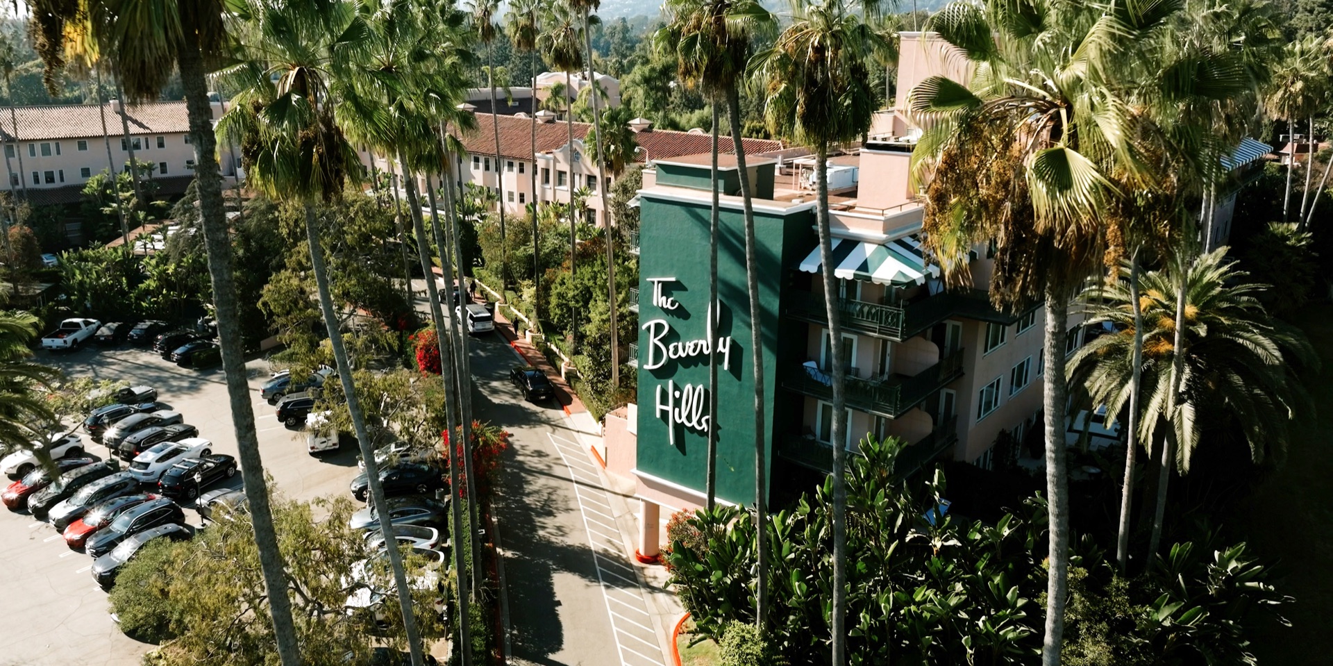 Aerial view of The Beverly Hills Hotel, its pink buildings and green script signage rising above the palm-lined drive in Beverly Hills, California