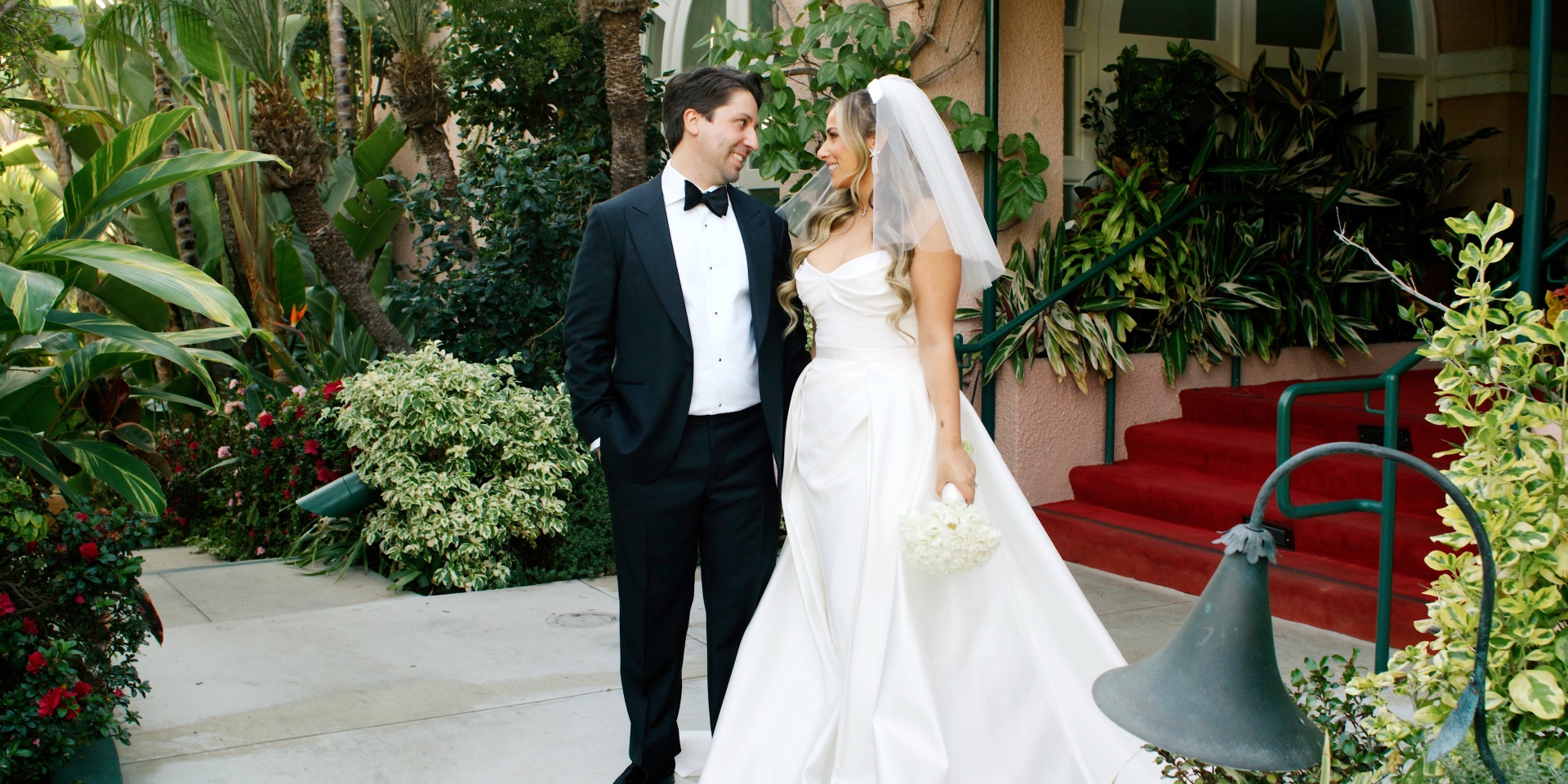 Bride and groom walking through The Beverly Hills Hotel garden past the red-carpeted steps and lush foliage