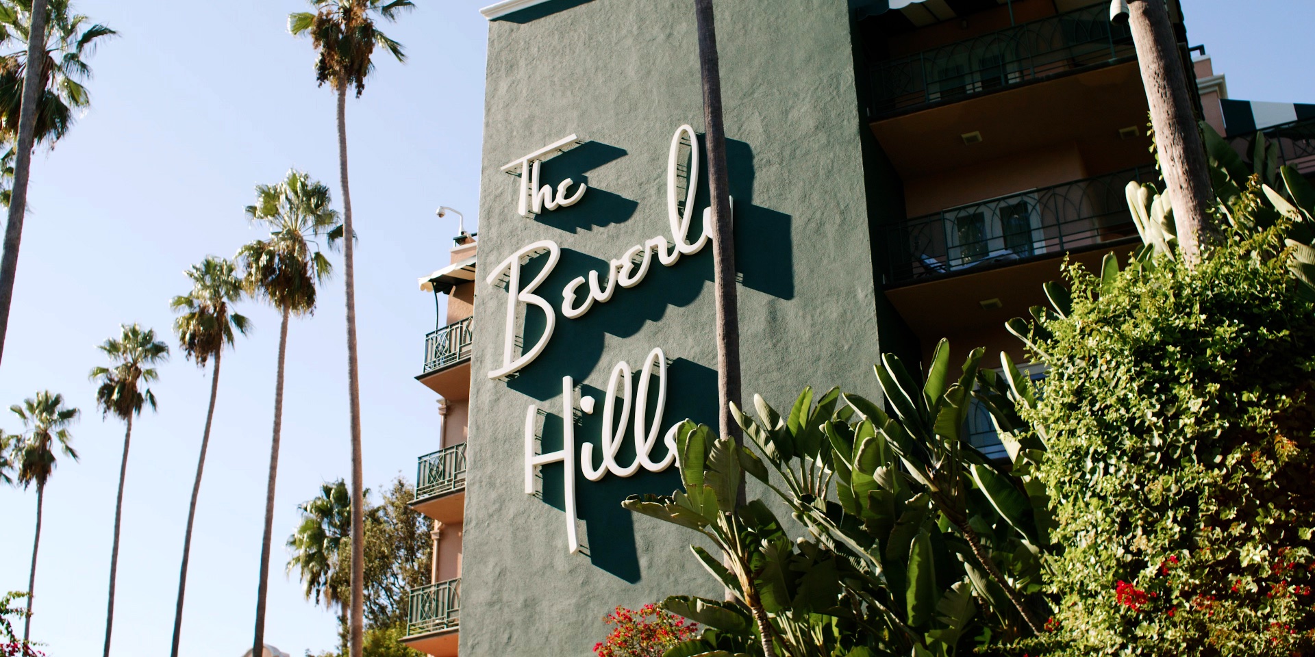 The Beverly Hills Hotel's iconic green and white script sign against tall palms and a clear blue sky