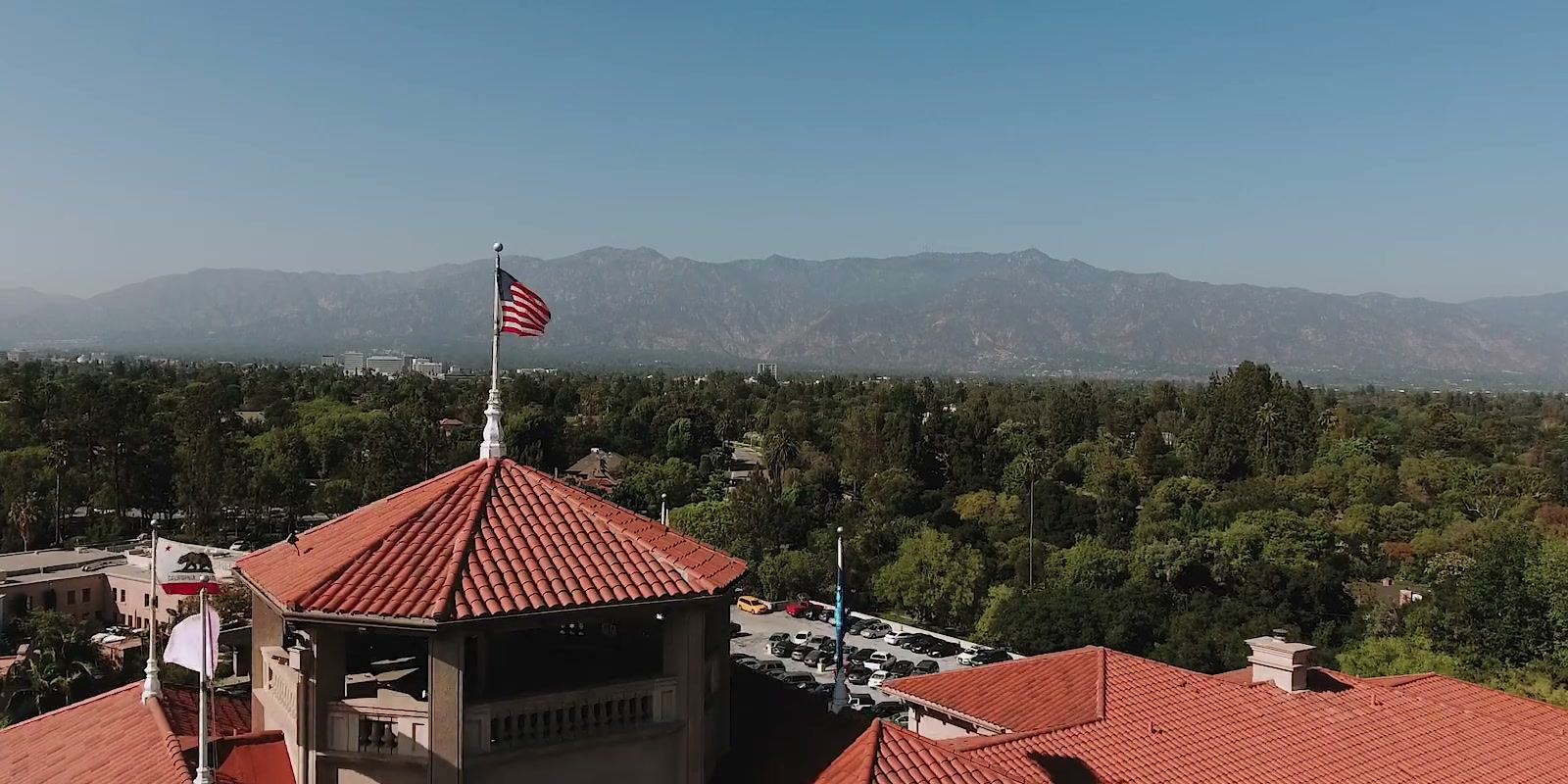 Red tile roofs and flags from above, with trees and the mountains beyond under a clear sky.