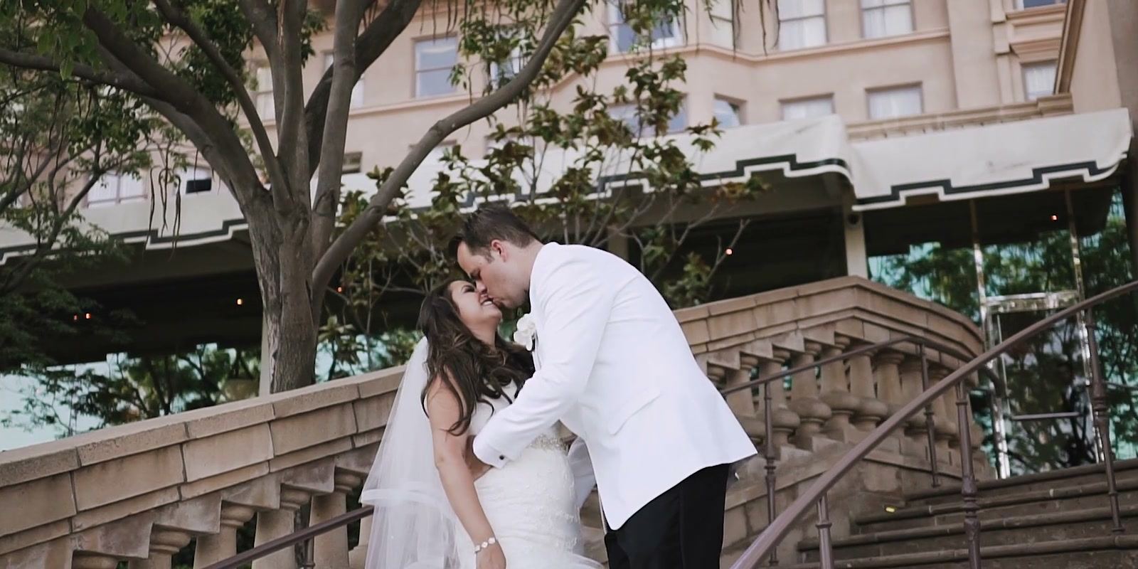 Alex leaning in to kiss Casey on the balustraded stone staircase below the hotel.