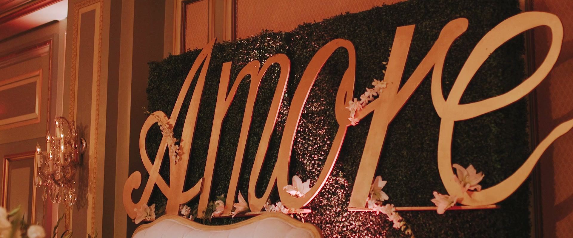 A gold script sign reading Amore set against a hedge wall in the ballroom, crystal sconces on the wall beside it.