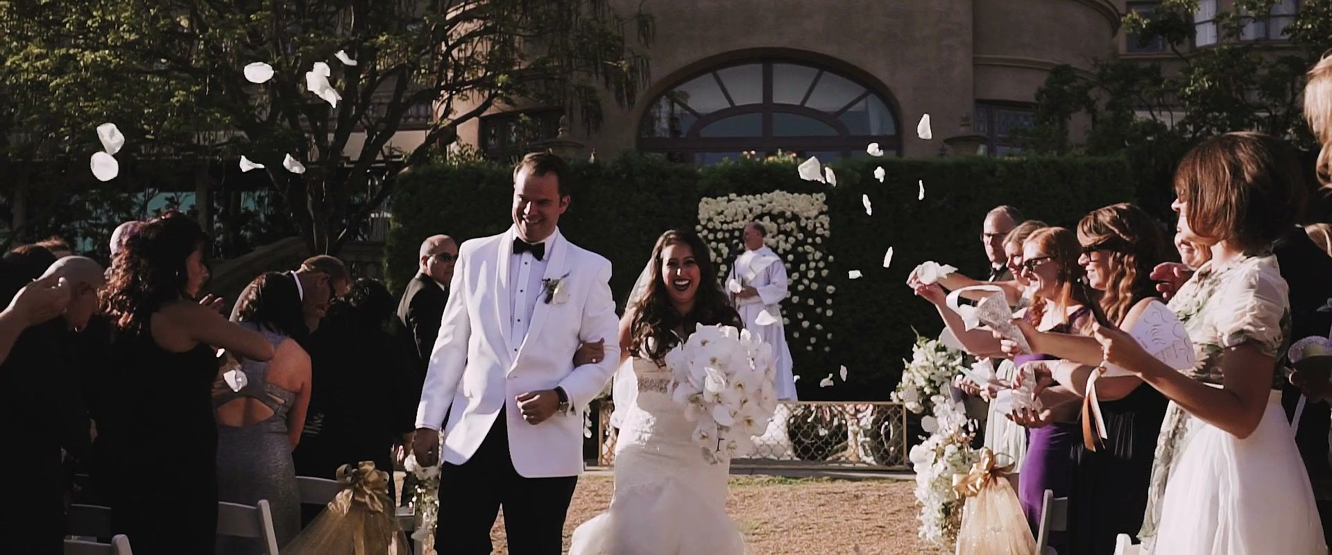 Casey and Alex walking back up the aisle through a shower of white petals, guests on both sides and the hotel’s arched window behind.