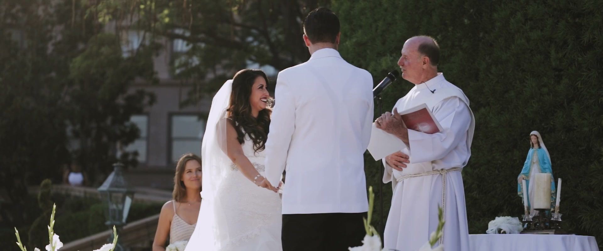 Casey smiling up at Alex as they hold hands for their vows, the officiant reading beside them and a statue of Mary on the table at right.