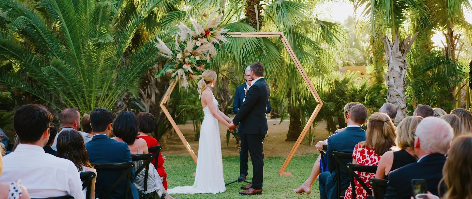 The ceremony beneath a hexagonal copper arch dressed with pampas grass, guests in black chairs on the lawn among the palms.
