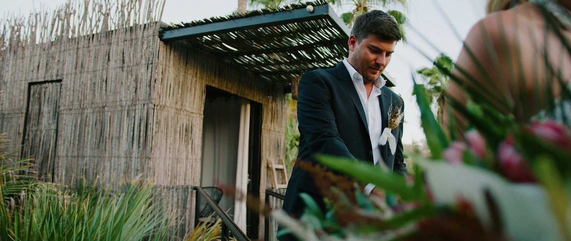 Cameron standing beside a reed-walled cabin, a bouquet soft in the foreground.