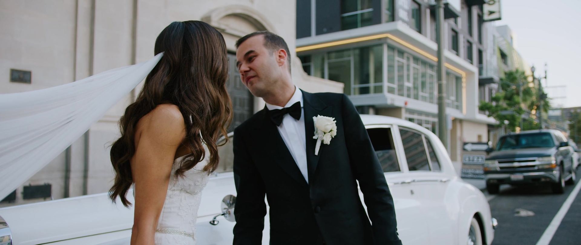 Desiree and Dominic on the sidewalk beside a white vintage car.