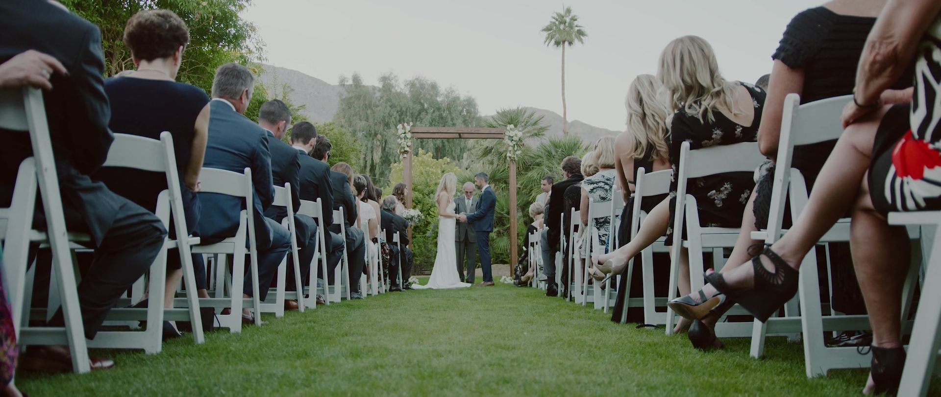 The aisle seen from the back, guests in white chairs on the lawn and the couple at the far end under the pergola.
