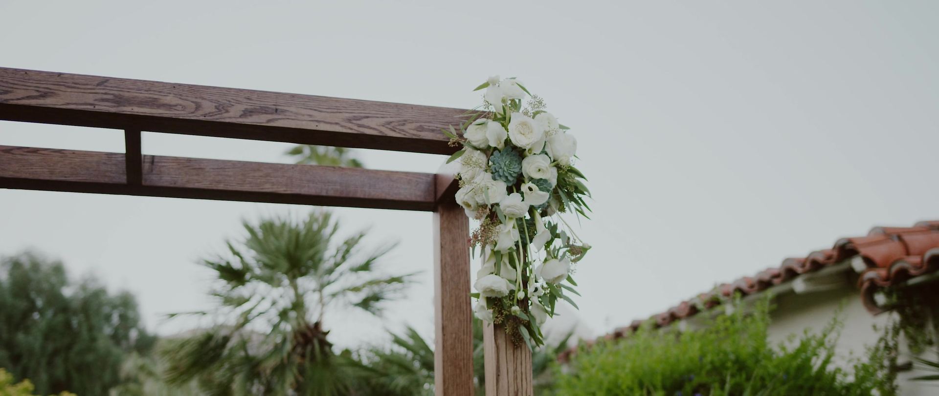 The ceremony pergola, a wooden frame dressed with white blooms against the palms and a tiled roof behind.