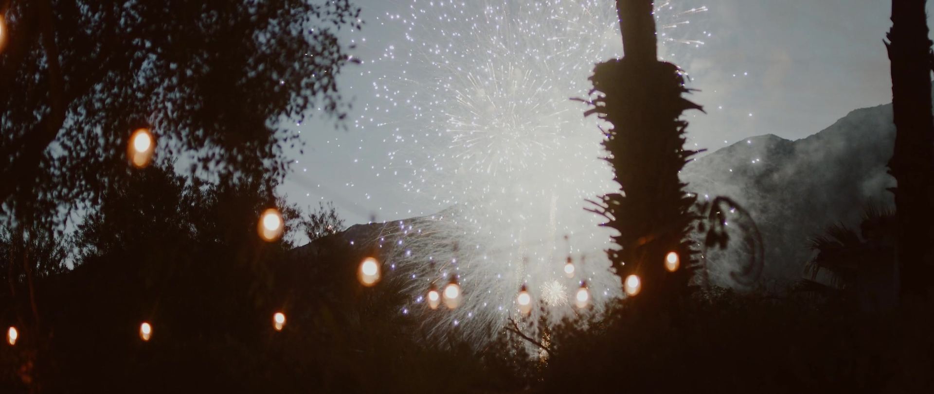 Fireworks bursting over the mountains beyond the string lights and palms, the sky still pale.