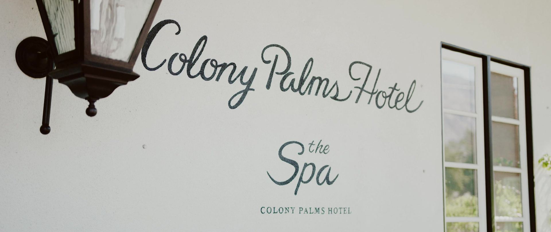 The painted Colony Palms Hotel sign on a white wall, a lantern beside it and a paned window to the right.