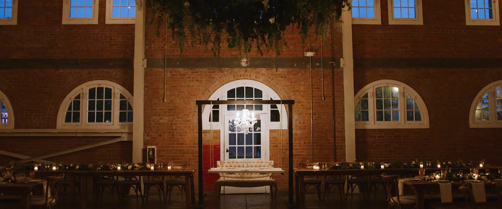 The brick hall set for the reception, arched windows, greenery hung from the beams, a chandelier above a white sofa and long wooden tables lit with candles.