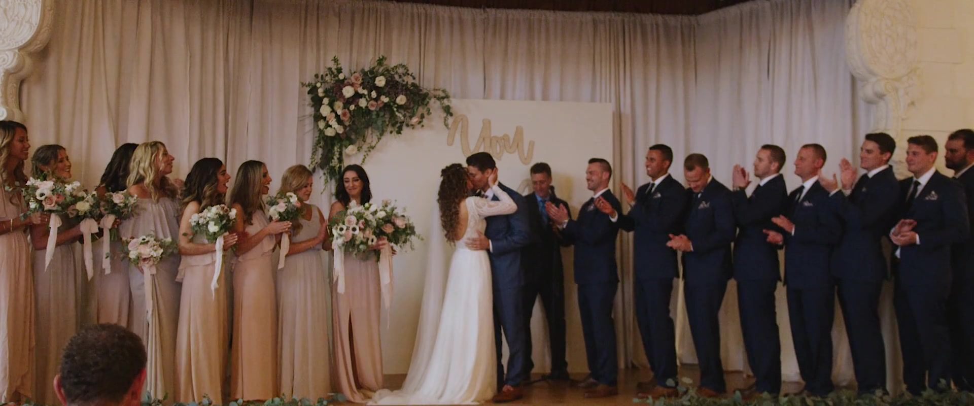 Dani and Shane kissing at the altar beneath a floral arrangement, bridesmaids in champagne to one side and groomsmen in blue applauding on the other.