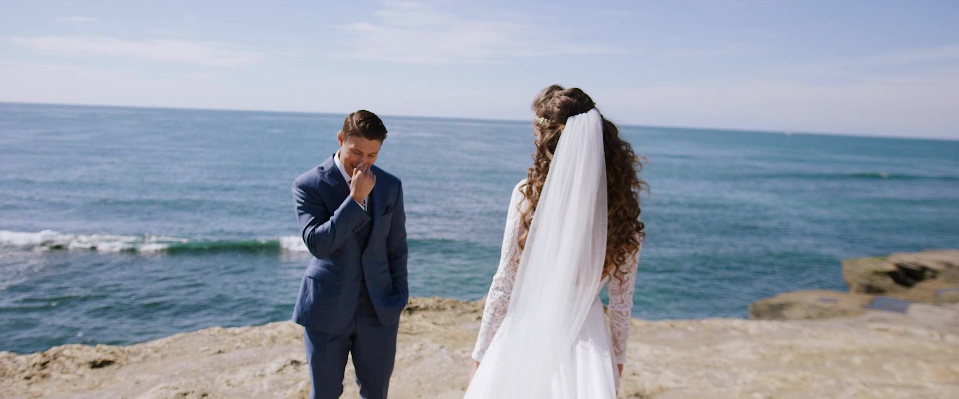 Shane with a hand to his mouth as Dani walks toward him across the rocks, her veil lifting, the ocean behind them.