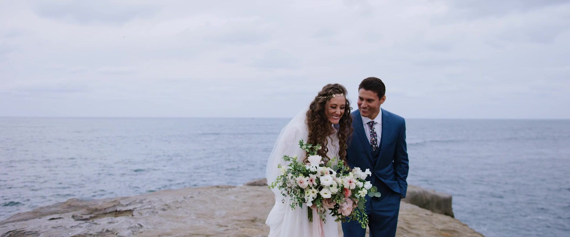 Dani and Shane on the rock ledge above the water, her bouquet of white and blush blooms held in front of her.