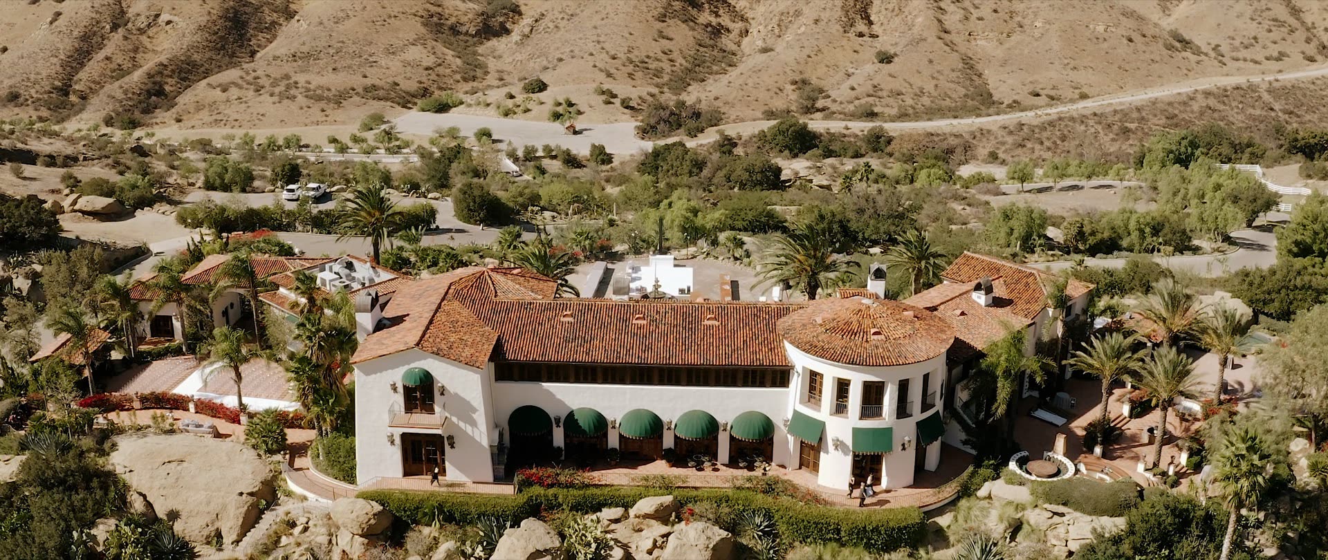 Daytime aerial of Hummingbird Nest Ranch, the white Spanish villa with terracotta roof and green colonnade awnings below dry Santa Susana hills.
