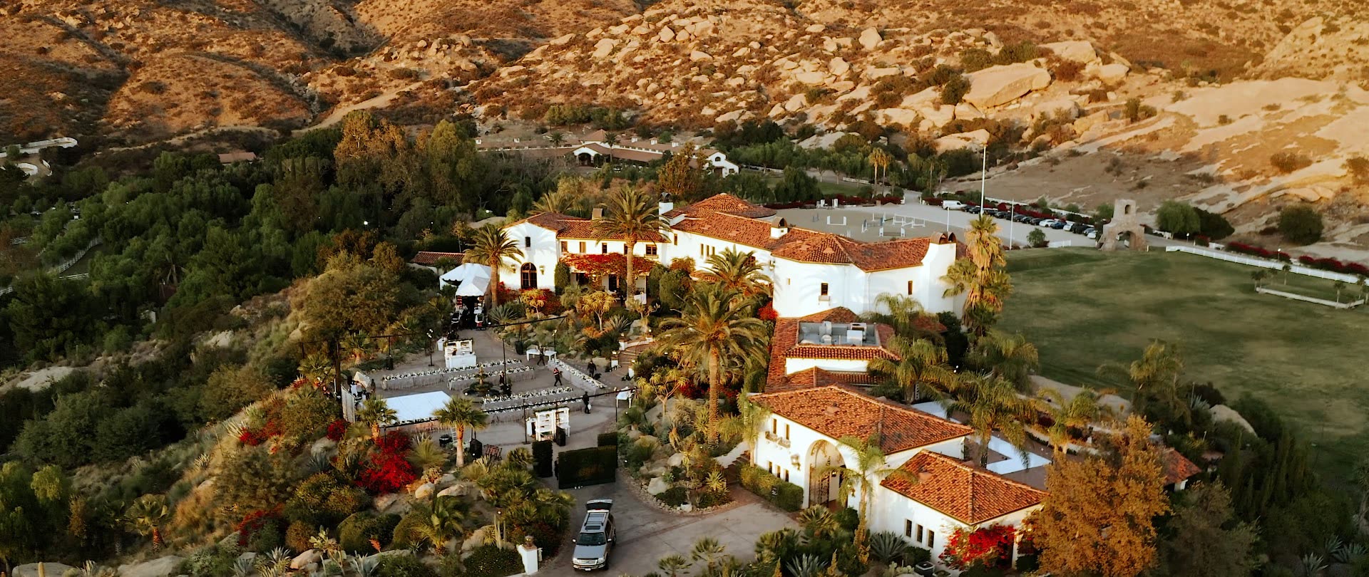 Golden-hour aerial of Hummingbird Nest Ranch, its Spanish hacienda, palm courtyards, and long candlelit reception table with the Santa Susana rocks glowing behind.