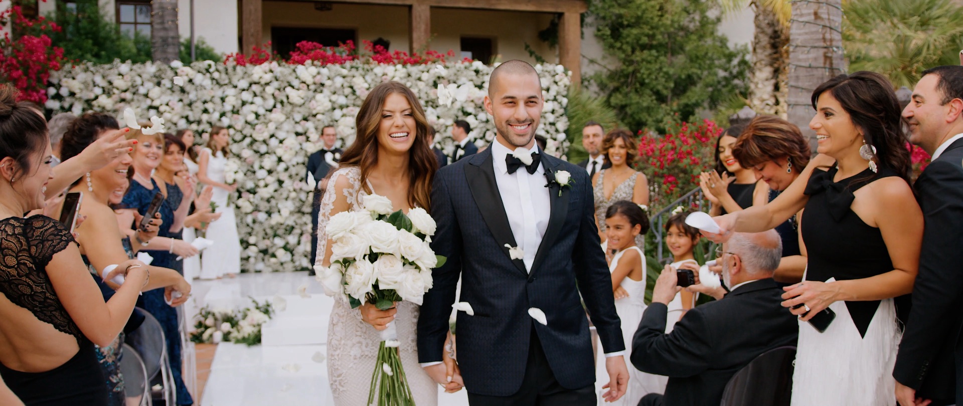 Edmond and Lauren walk their recessional through tossed white petals past a blush and white rose wall at Hummingbird Nest Ranch.