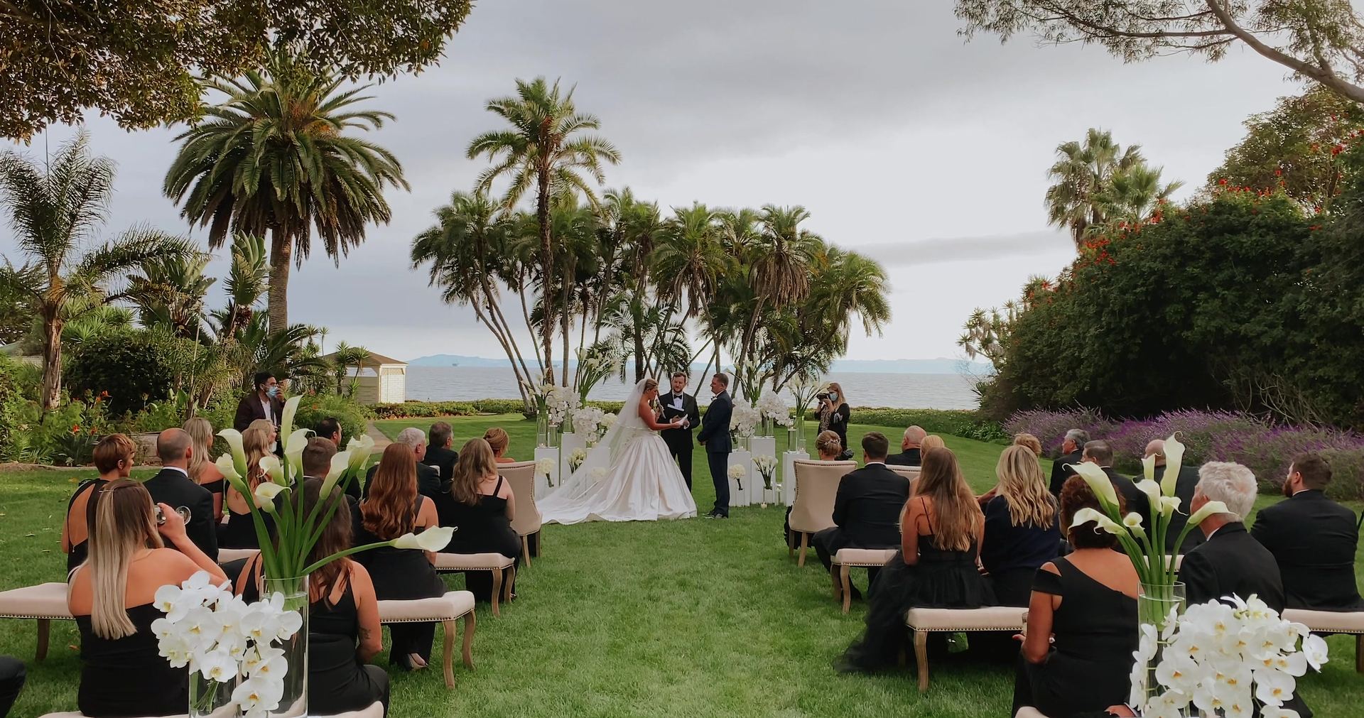 The ceremony on the lawn, guests on benches among the palms with the ocean and an island beyond.