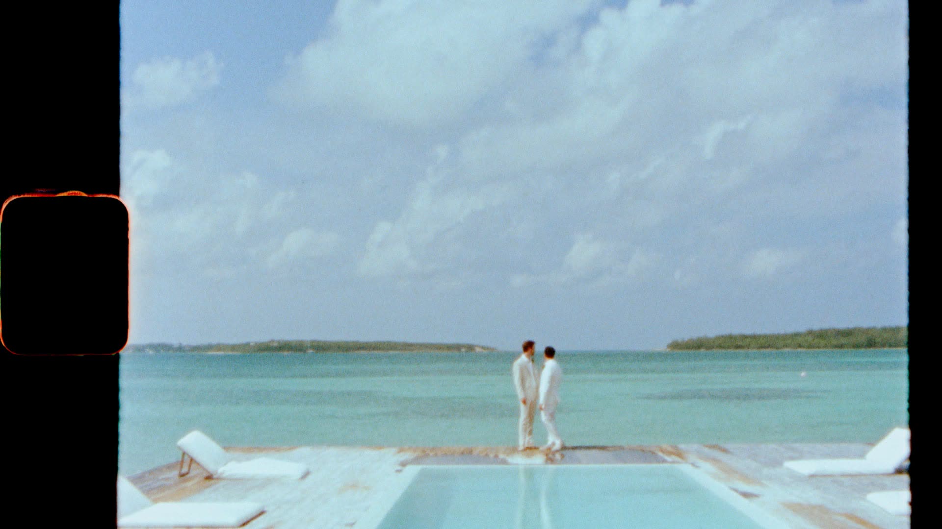 Izak and Slater in white at the far end of a pale deck running out over flat turquoise water, a low green cay on the horizon, at their private island wedding in the Bahamas