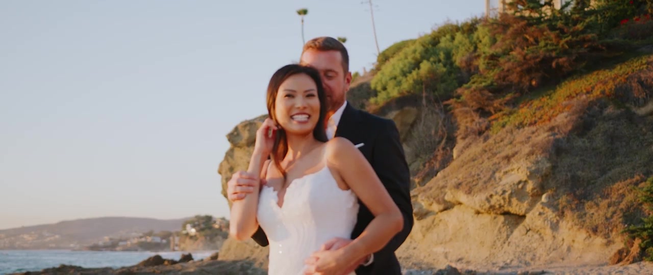 Jan smiling in her wedding dress as Brian holds her from behind on the sand at Montage Laguna Beach, the bluff and warm evening light behind them