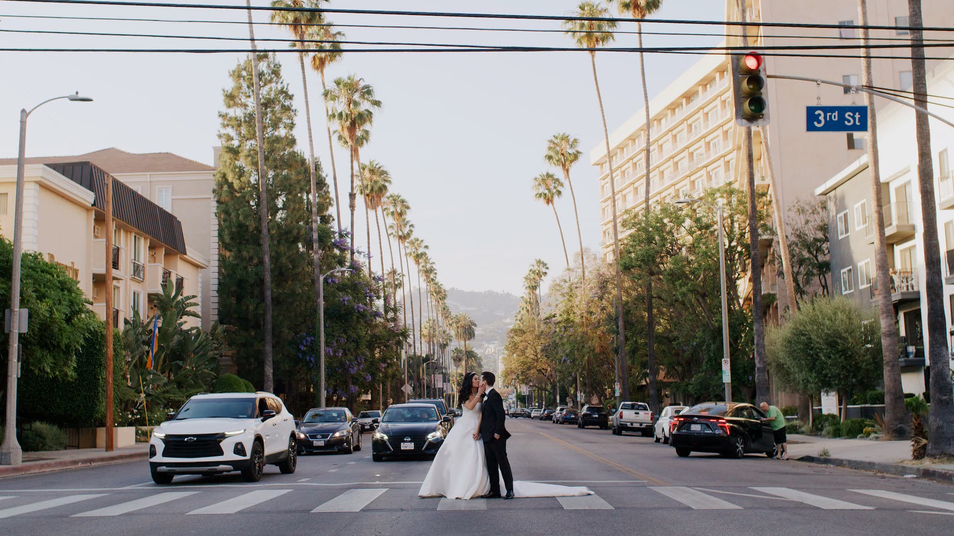 Joe and Jessica standing together in a crosswalk on a wide palm lined street near Four Seasons Hotel Los Angeles at Beverly Hills, a traffic light and a 3rd Street sign behind them