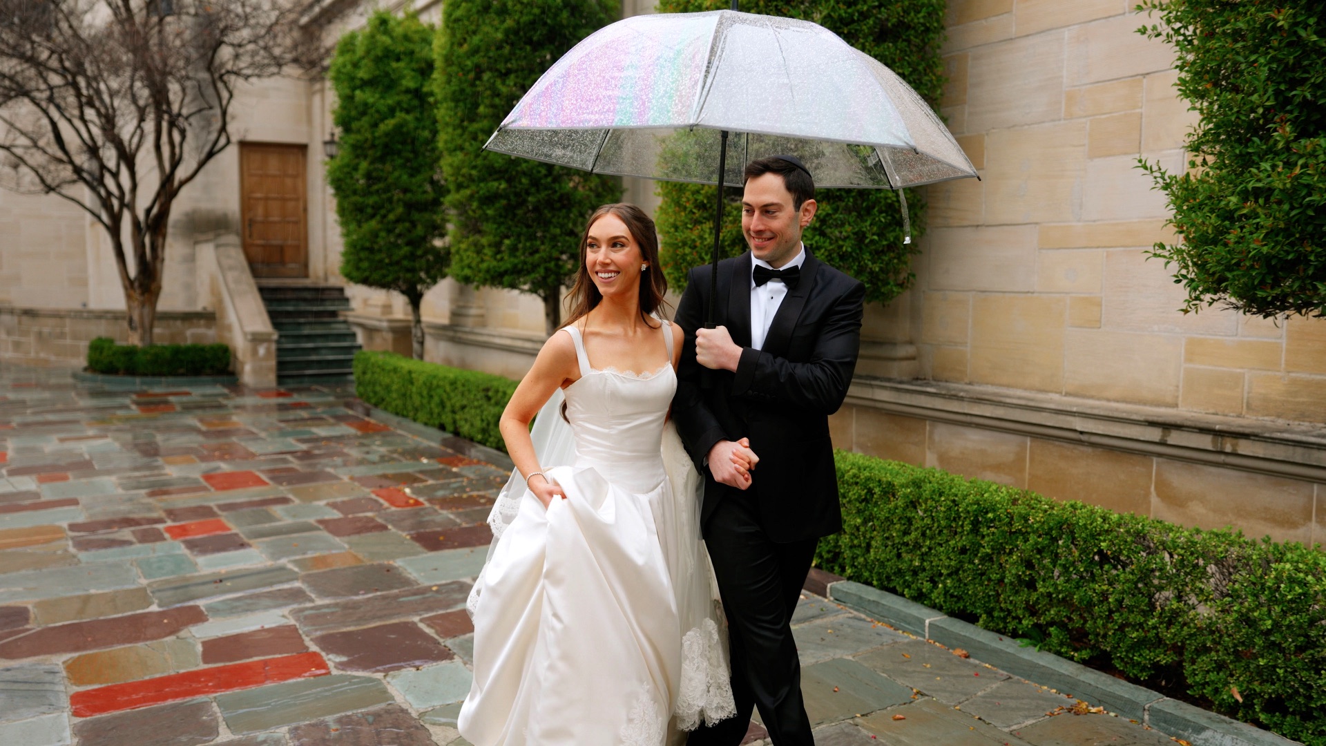 Jonathan and Alexa walking under a clear umbrella in the rain on their Beverly Hills wedding day