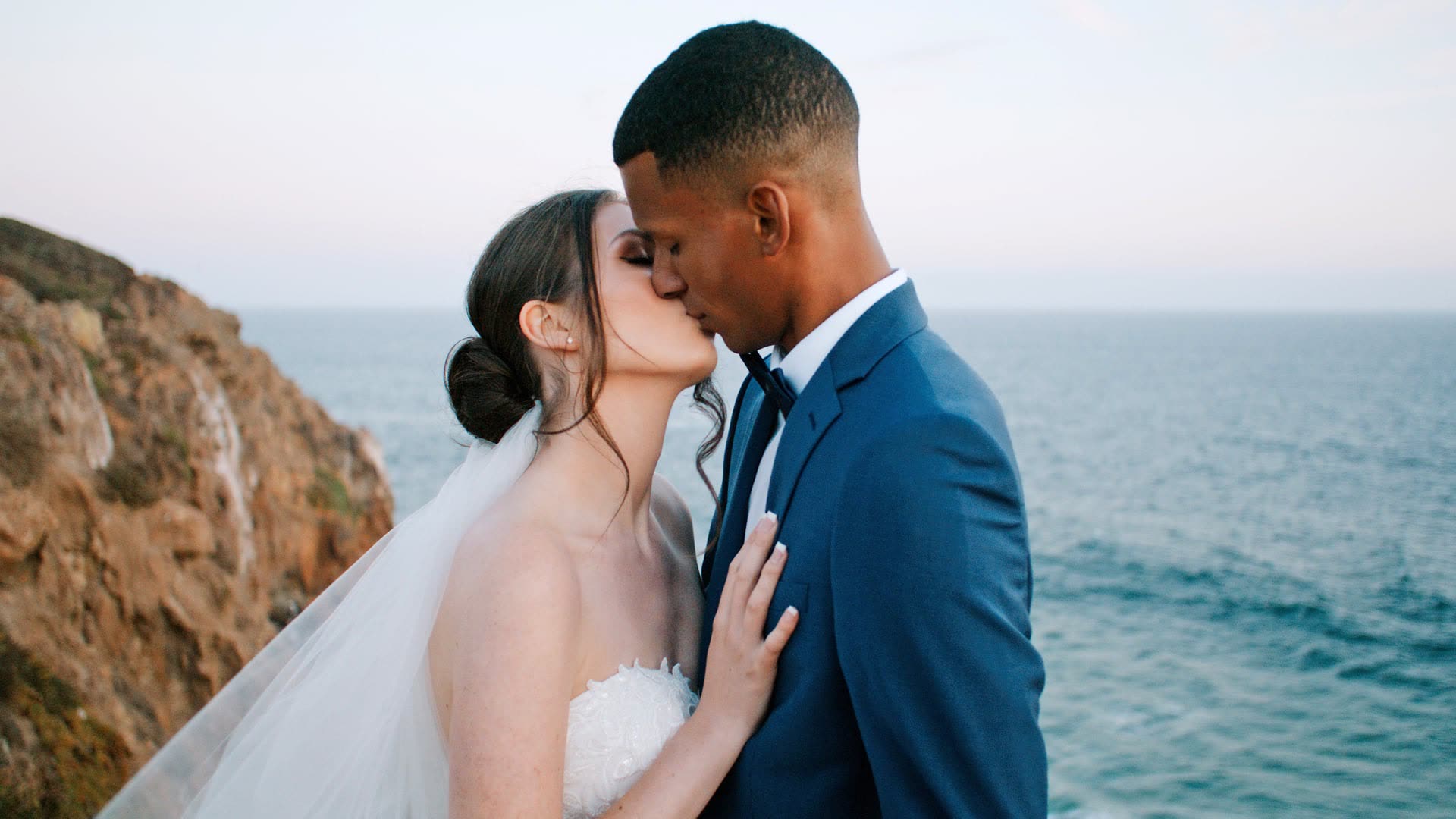 Lucas and Lindsey kissing on the San Diego coast, open ocean and a sandstone cliff face behind them