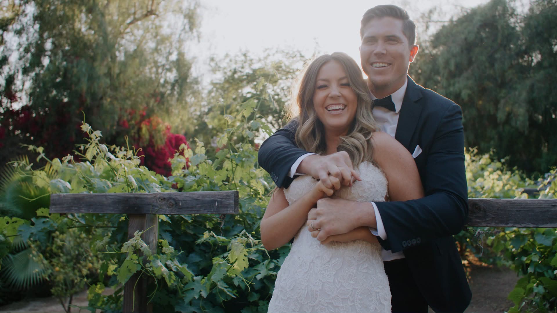 Shane and Rosie standing among grape vines beside a rustic split-rail fence in Riverside, his arms around her from behind, bougainvillea and greenery around them.