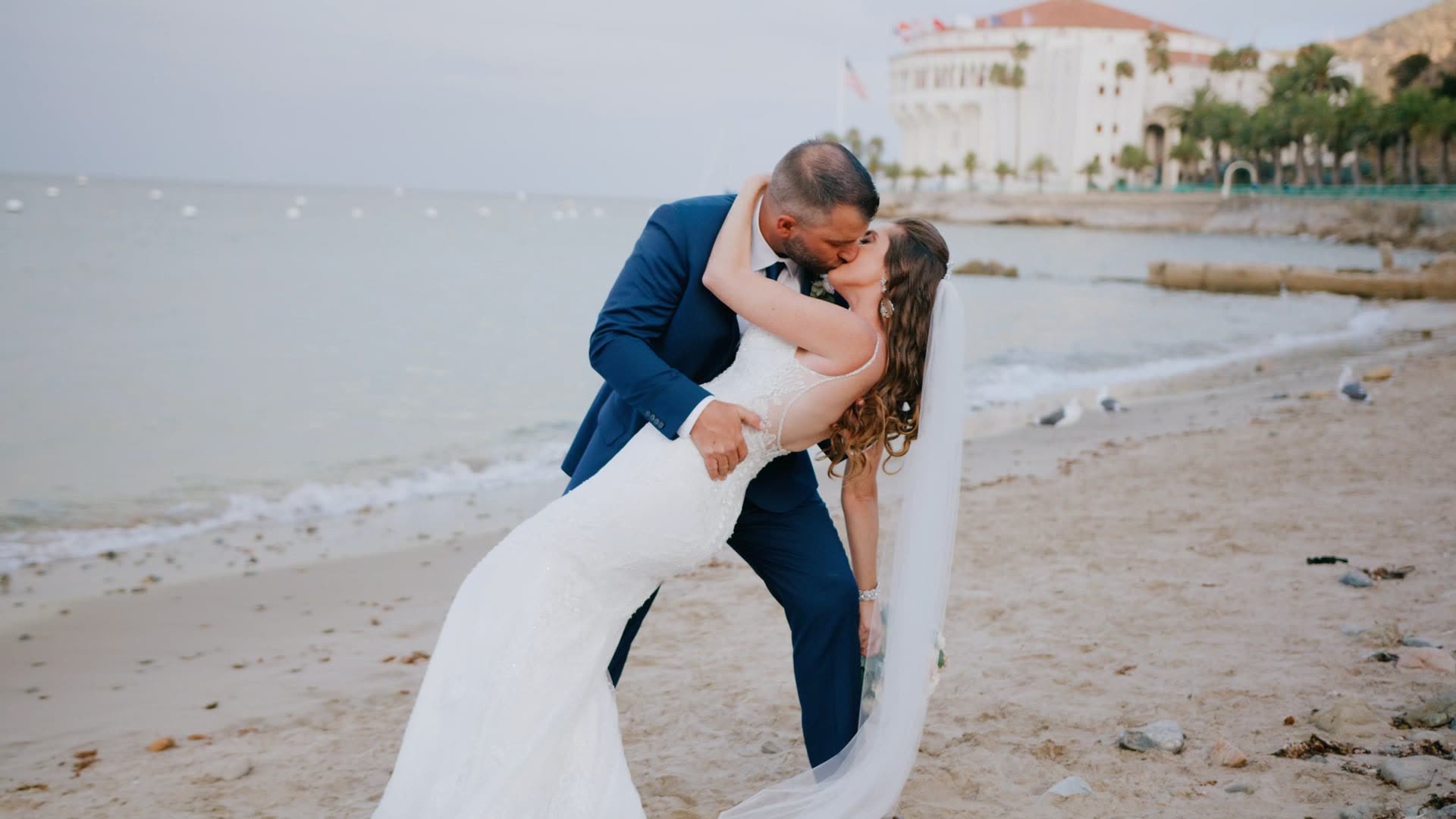 Timothy and Amber in a dip kiss at the water's edge on the sand in Avalon, the tiled waterfront buildings of Catalina Island behind them.
