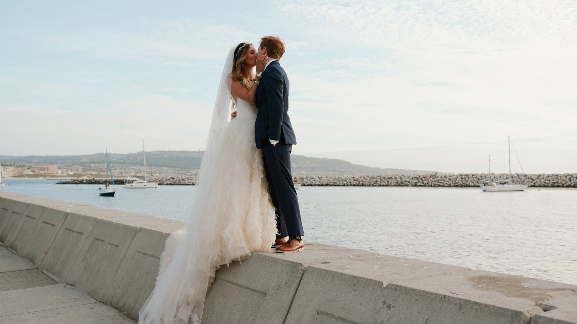 Tyler and Rachel standing on the concrete seawall above King Harbor at The Portofino Hotel and Marina in Redondo Beach, moored boats and the rock breakwater behind them in low late light