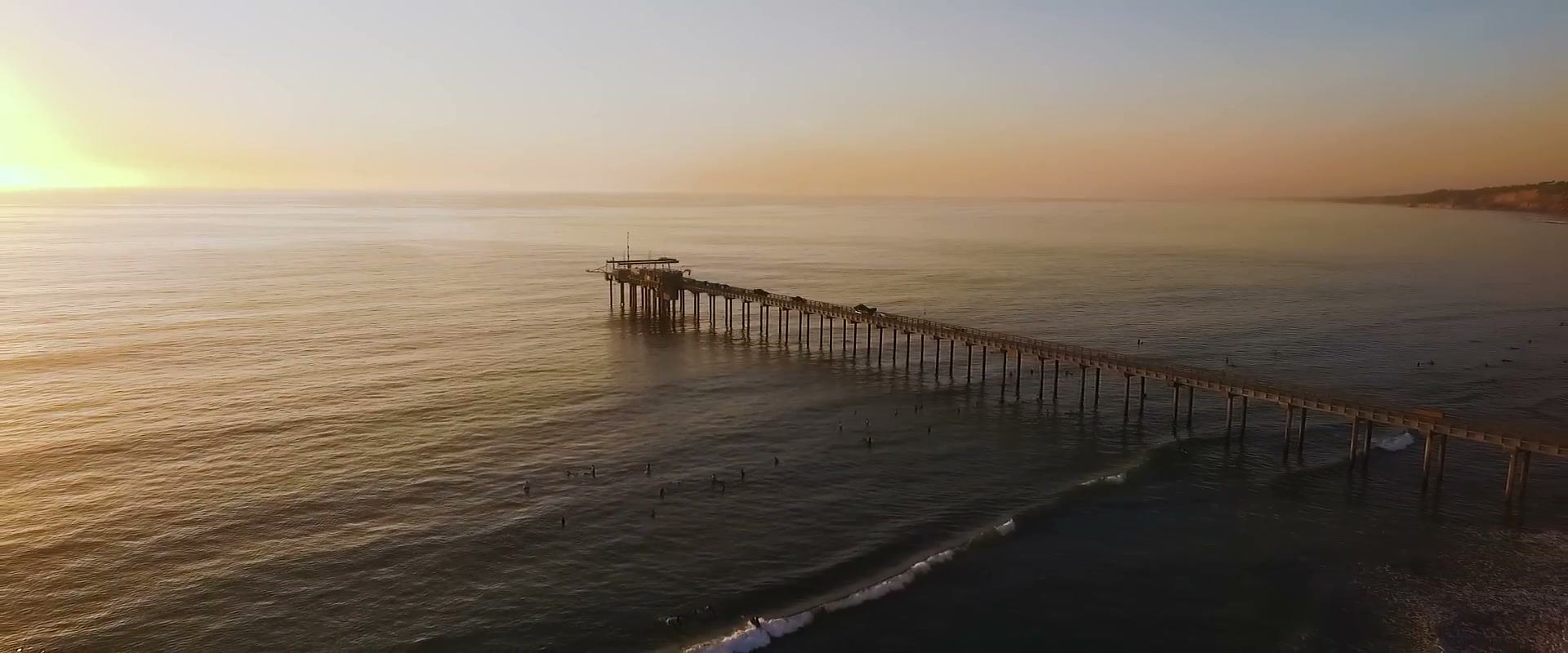 Scripps Pier from above, reaching out across a calm ocean with the low sun at the left edge of the frame and surfers in the water beside the pier.