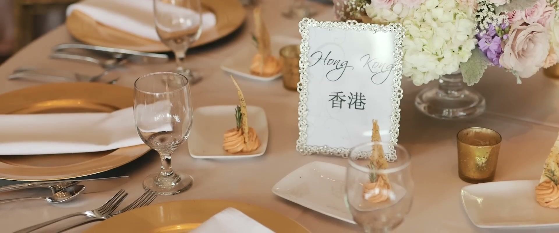 A reception table set with gold chargers, small plates and a silver-framed table card reading Hong Kong beside pale roses and hydrangea.