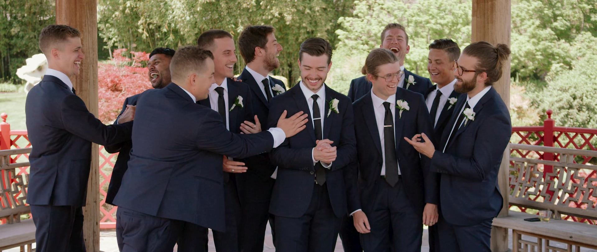 The groomsmen together in a wooden pavilion with red lattice railings, maples and a pond behind.