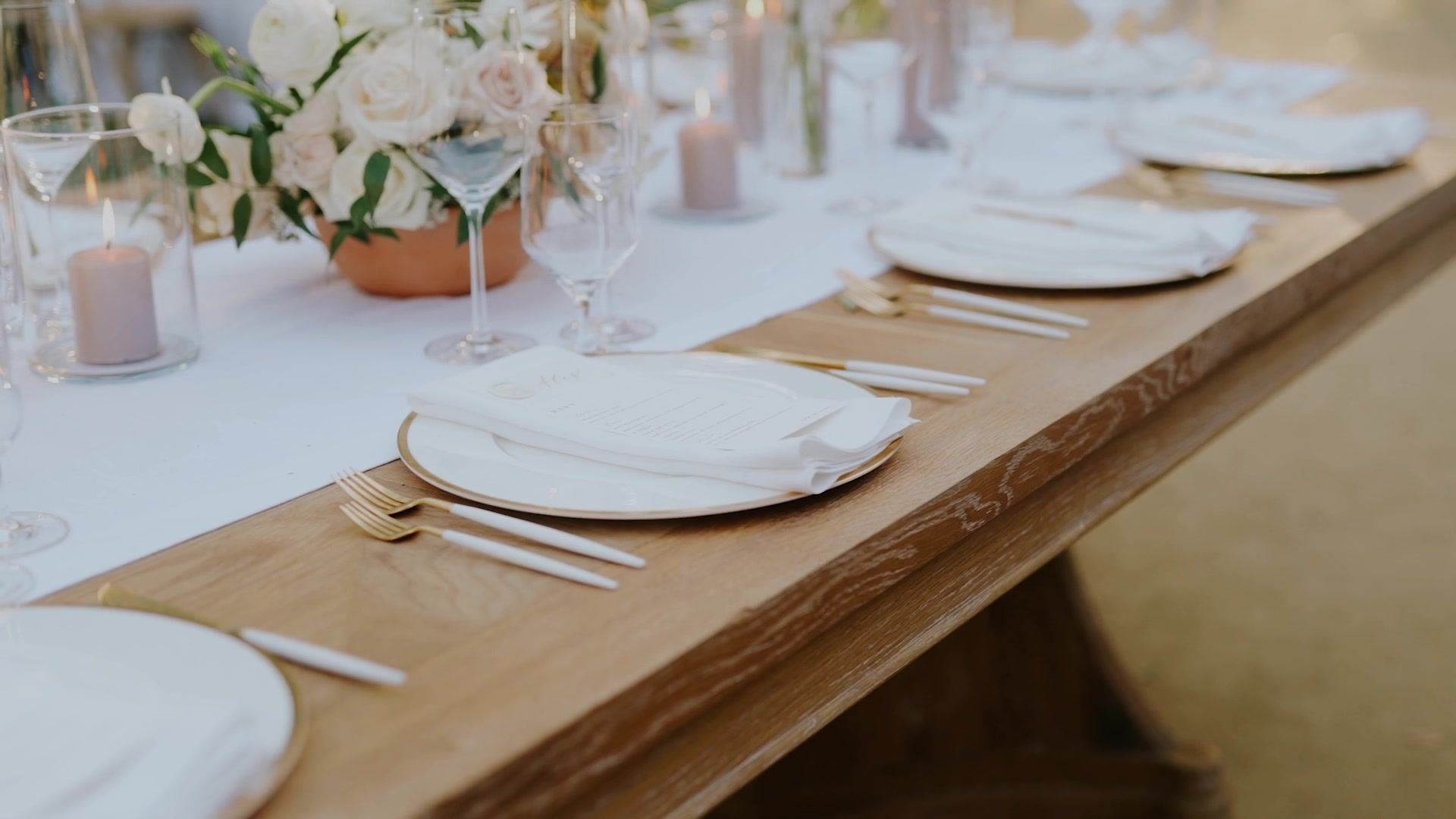 The wood reception table with a white runner down its centre, blush pillar candles in glass and a low arrangement of white and blush roses in a copper bowl.