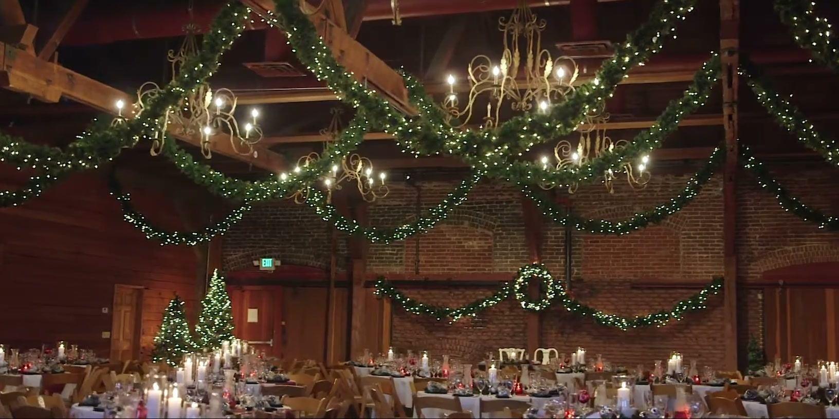 The main hall strung with lit pine garland and a wreath, gilt chandeliers and two Christmas trees above round candlelit tables.